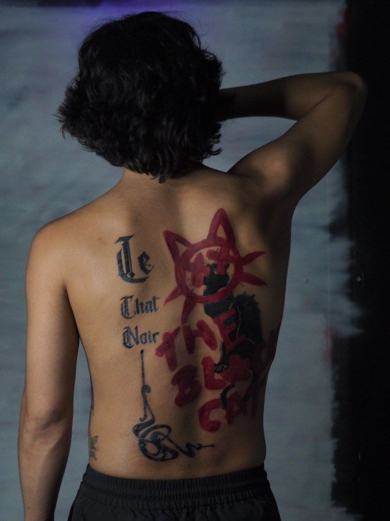 A large tattoo on the back. Written in gothic text sitting vertically down the left side of the back are the words ‘le chat noir’ with an art deco style corner border embellishment framing the image sitting next to the text. The tattoo which is to the right of the text is a cartoonish drawing of a cats face in thick red lines that look like they were painted on with a big paint brush. It is a circle with two pointy ears two whiskers on each side whiskers and has crosses as eyes. There is text beneath it in the same thick red lines and childlike style that reads ‘the black cat’ Behind the red design is an all black sitting and looking out at the viewer as if from behind a red gate.