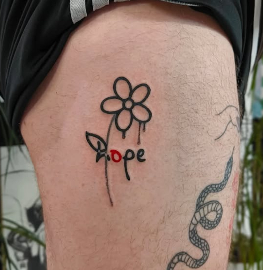 A simple cartoon style daisy in the upper thigh in thick black lines. The stem is part of a previous design which the flower is covering up, text crosses the middle of the flower with the same bold lines and casual hand written style. The first letter is a lowercase h which overlaps the leaf and stem. The next letter is o which is tattooed in red ink, followed by letters p and e to spell out hope.