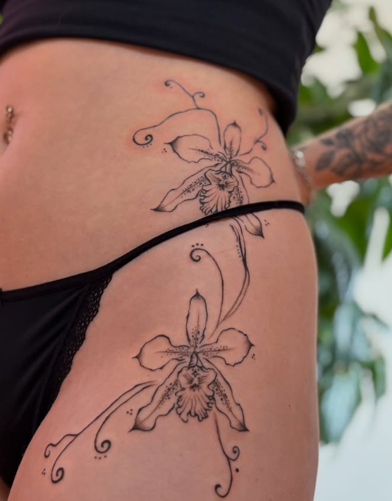A black line work tattoo with some minimal shading and dot work. The design depicts two blooming orchids cascading down the hip to the upper thigh with tendrils that come away from the flowers in curls.