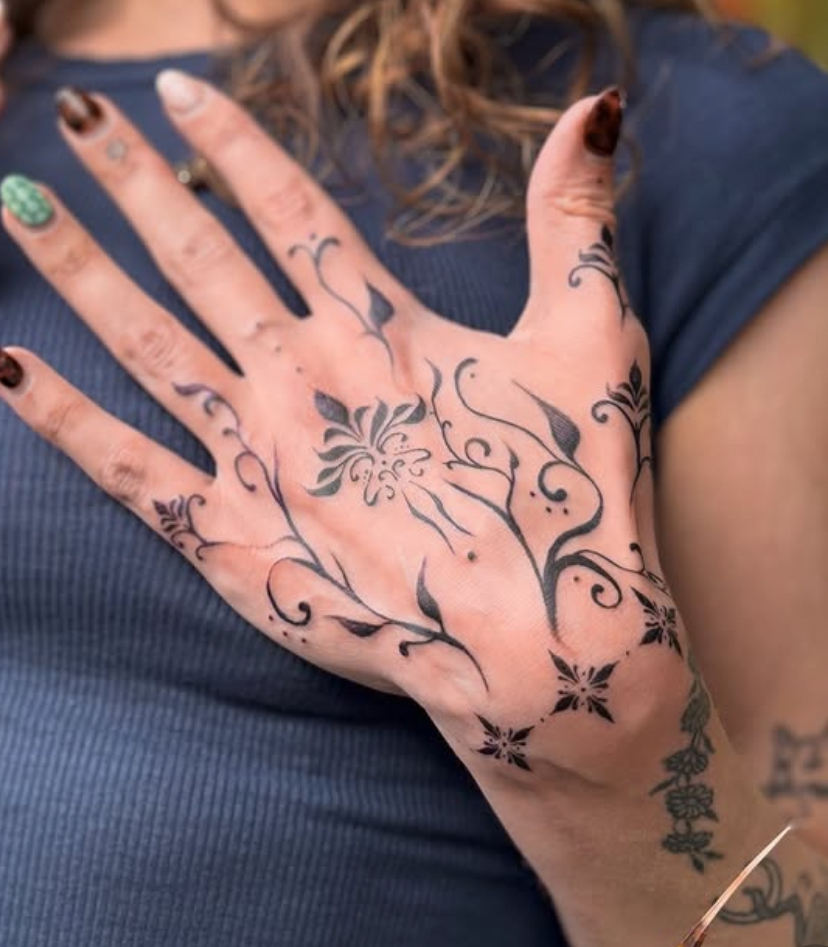 Ornamental tattoos on the back of the hand in black ink. There are dots and swirly plant like patterns on two sides, with art nouveau flowers similar to orchids In the middle of the hand and up the thumb. There are three simple flowers at the bottom of the hand and top of the wrist that act like a border to the work.