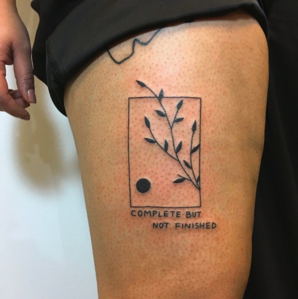 A black linework tattoo on the middle of the thigh, the design is within a black rectangular frame with a small black circle in the lower left corner, there is a simple branch with leaves coming from the inside right corner and spreading diagonally and upwards across the rectangle, the tip of the branch breaks free from the rigid shape and is growing upwards and to the left. There is staggered text beneath the rectangle in a simple handwritten style that says ‘complete but not finished’