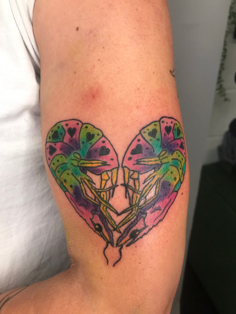 A tattoo on the inner arm of two multicoloured shrimps - hot pink, lime green, turquoise, purple and yellow- embracing to make the shape of a heart. The shrimps are outlined in black and decorated with little black love hearts on their shells