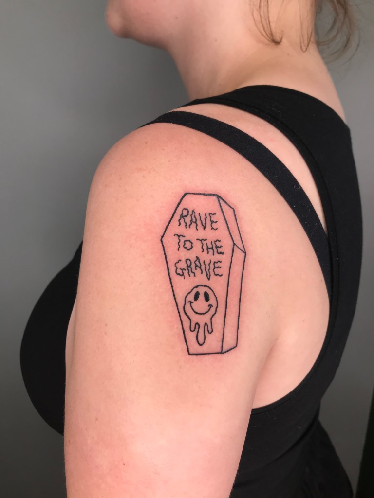 A tattoo on the shoulder in the shape of a coffin, on the lid there is wobbly text which says ‘rave to the grave’ with a wobbly smily face beneath it. The tattoo is done with simple black line work and no shading.